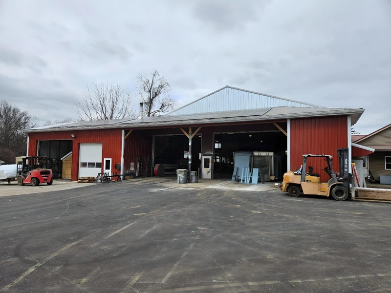 Red barn-style manufacturing workshop with open bay doors, forklifts, and building equipment